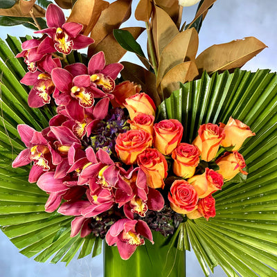 A Dream Come True arrangement with orange roses, cymbidium orchids, and fan palms in a tall glass vase, same-day delivery by Santa Monica Florist.