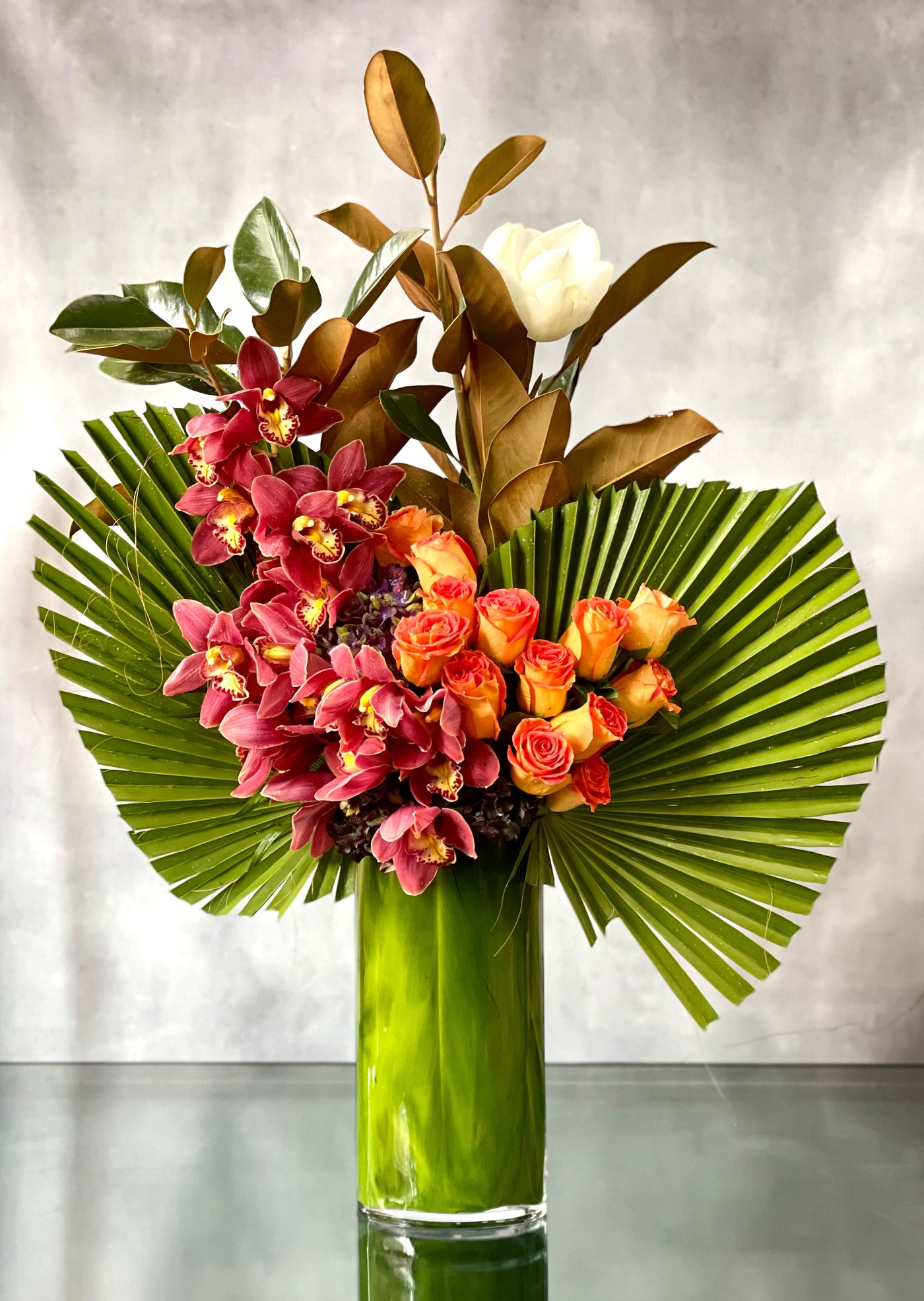 A Dream Come True arrangement with orange roses, cymbidium orchids, and fan palms in a tall glass vase, same-day delivery by Santa Monica Florist.