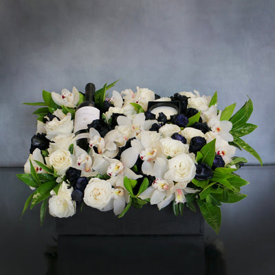 A Gift For You arrangement with black and white roses, cymbidium orchids, greens in a black wooden box with wine and a candle, by Santa Monica Florist.