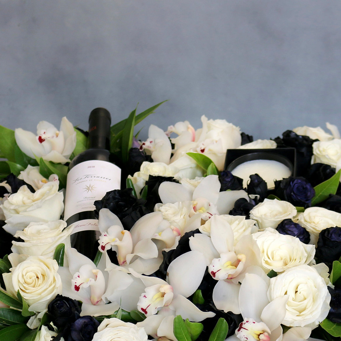 A Gift For You arrangement with black and white roses, cymbidium orchids, greens in a black wooden box with wine and a candle, by Santa Monica Florist.