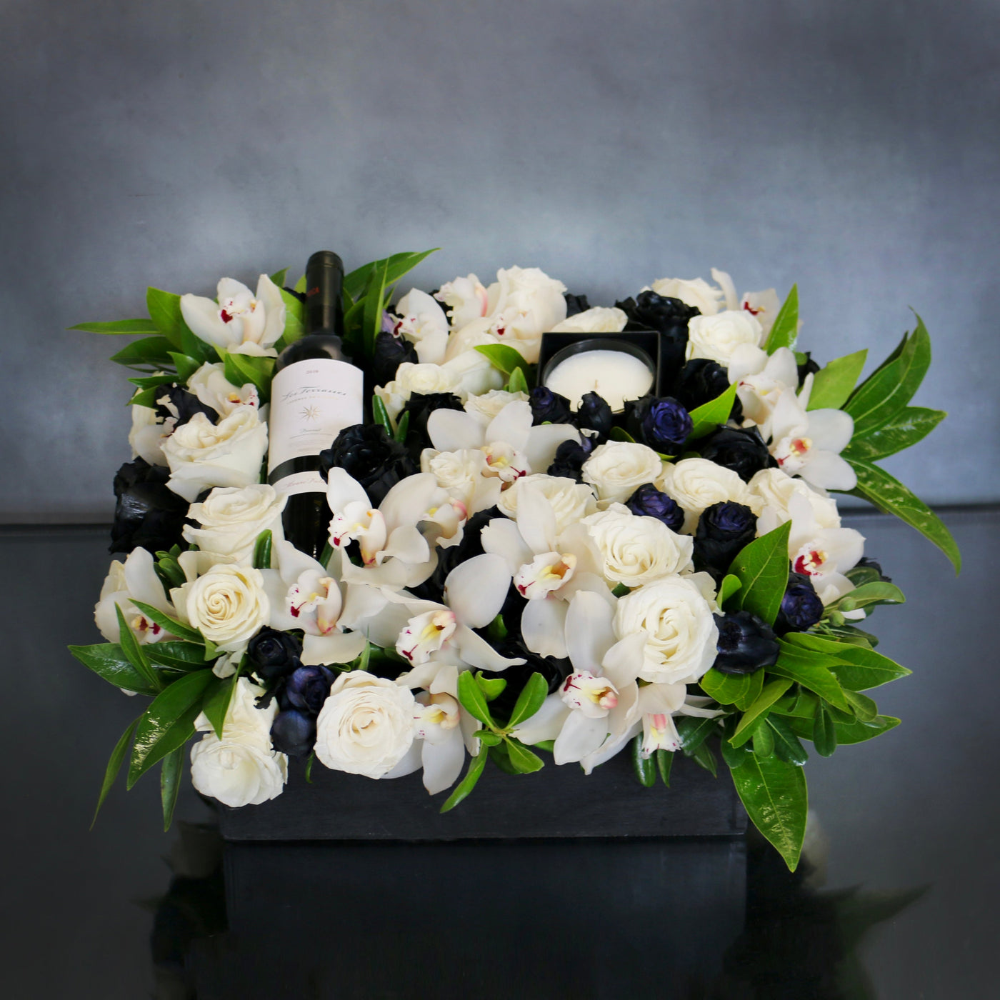 A Gift For You arrangement with black and white roses, cymbidium orchids, greens in a black wooden box with wine and a candle, by Santa Monica Florist.