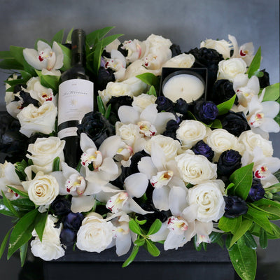 A Gift For You arrangement with black and white roses, cymbidium orchids, greens in a black wooden box with wine and a candle, by Santa Monica Florist.