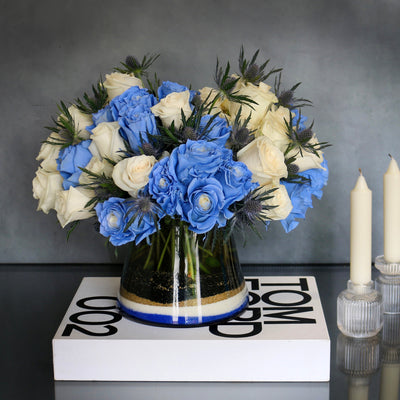 Blue Me Away arrangement with blue and white roses, thistles, and colored sand in a modern vase, available for same-day delivery by Santa Monica Florist.