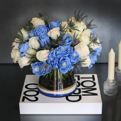 Blue Me Away arrangement with blue and white roses, thistles, and colored sand in a modern vase, available for same-day delivery by Santa Monica Florist.