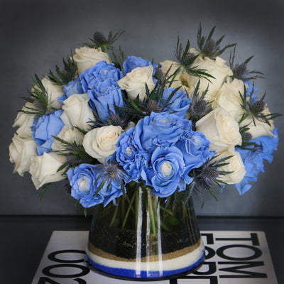 Blue Me Away arrangement with blue and white roses, thistles, and colored sand in a modern vase, available for same-day delivery by Santa Monica Florist.