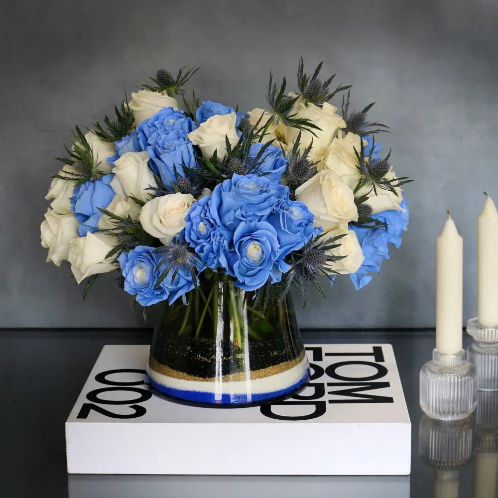 Blue Me Away arrangement with blue and white roses, thistles, and colored sand in a modern vase.
