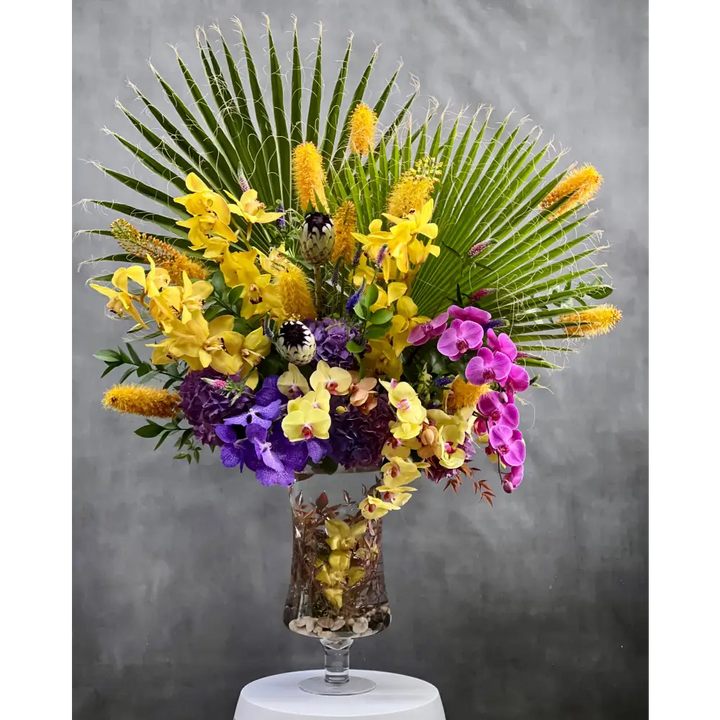 A Spectacular Floral Fantasy featuring extravagant and exotic blooms in tropical colors on a grand scale by LA Orchidia for occasions like congrats, new baby, just because, love and romance. Vibrant seasonal blooms paint the oranges, deep pinks, purples, lime greens and yellow orchids and other floral. Large tropical leaves lined vase with this amazing design and colors. It's a one-of-a-kind, dynamic presentation that will be the center of attention wherever it is placed. Approx: 16 W x 30" H.