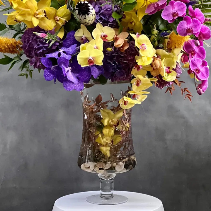 A Spectacular Floral Fantasy featuring extravagant and exotic blooms in tropical colors on a grand scale by LA Orchidia for occasions like congrats, new baby, just because, love and romance. Vibrant seasonal blooms paint the oranges, deep pinks, purples, lime greens and yellow orchids and other floral. Large tropical leaves lined vase with this amazing design and colors. It's a one-of-a-kind, dynamic presentation that will be the center of attention wherever it is placed. Approx: 16 W x 30" H.
