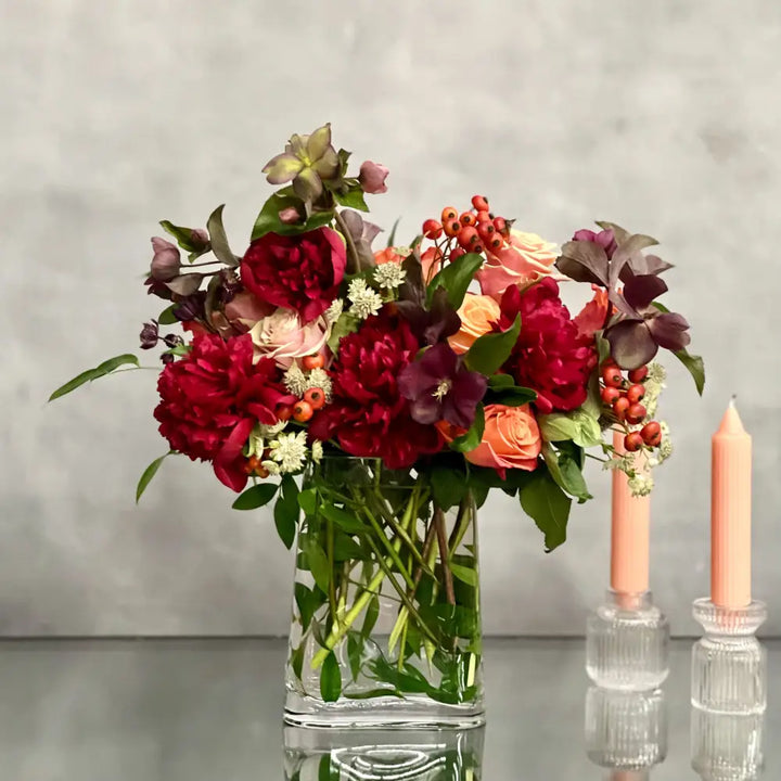 Majestic winter arrangement with orange roses, burgundy peonies, and winter accents in a glass vase.