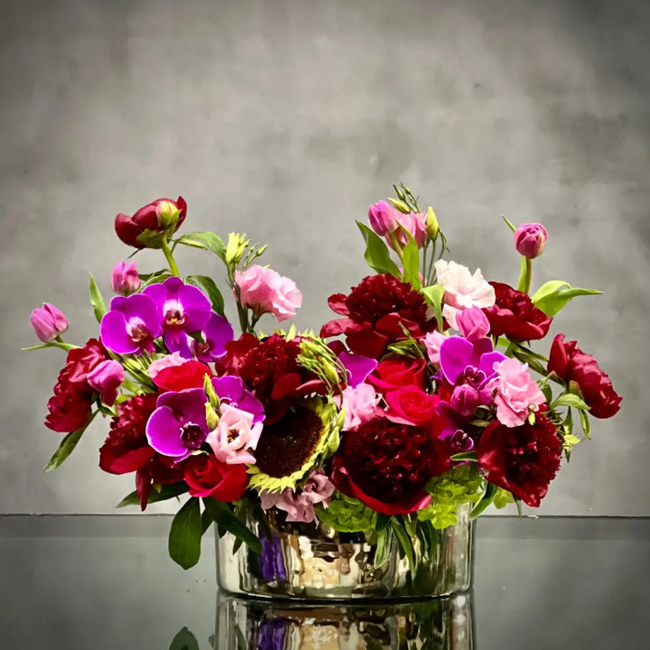 Luxurious Burgundy peony, lavender tulip, and purple orchid arrangement in a silver container.